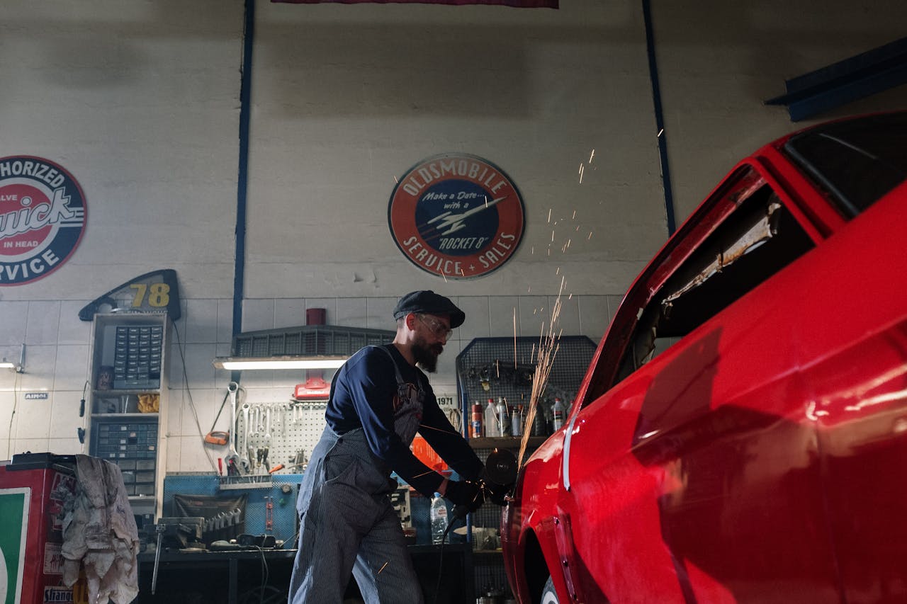 A bearded auto mechanic using a grinder on a red car in a vintage garage setting.